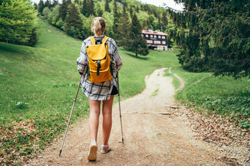 Lonely female with yellow backpack walking by mountain path with trekking poles to mount refuge hut...