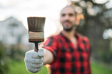 A young man in a work plaid shirt with a paintbrush in the backlit garden. The brush is in focus in the foreground.
