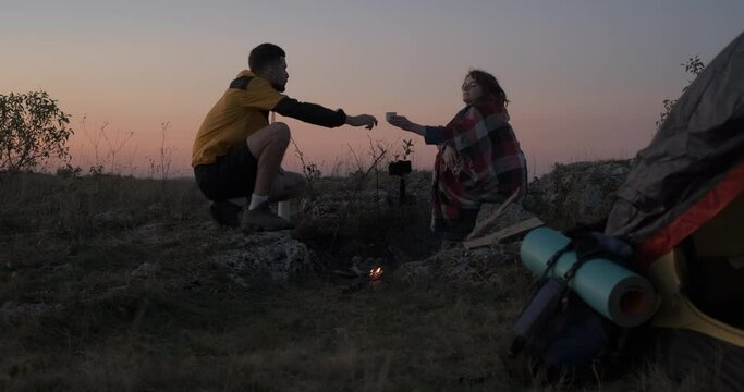 Two Young Travelers Man and Woman Sitting Near the Campfire and Tent Outdoors During Sunset and Drinking Hot Tea from Thermos