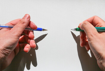 two hands of an ambidextrous young man holding a blue pen in his left hand and a green one in his...