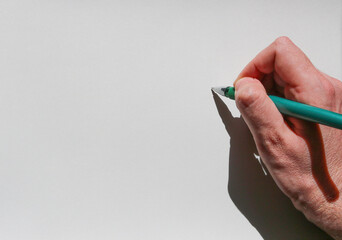 right hand of a young man holding a green pen tipped over a sheet of paper, with a shadow cast at sunset, and blank space to place a text or image