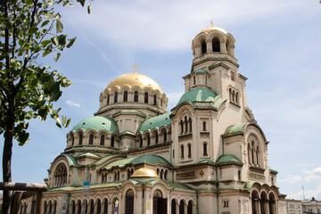 Fototapeta na wymiar Alexander Nevsky facade cathedral in the center of Sofia, capital city of Bulgaria, on a sunny day, with a tree and blue sky for a tourist walk - beautiful orthodox symbol for a travel around balkans