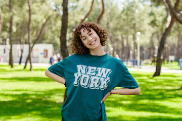 Young redhead woman wearing green tee standing on green city park, outdoors holding hands on hips and looking at camera with big smiles, closed eyes. Smiling confidence.