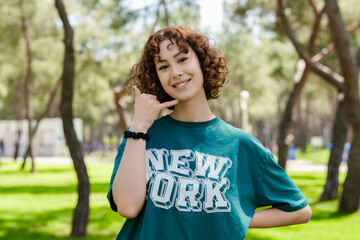 Naklejka premium Young redhead woman wearing green t-shirt standing on city park, outdoors smiling doing phone gesture with hand and fingers like talking on the telephone. Communicating concepts.