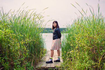 a brunette girl in a dress and a black jacket stands on the river bank on a wooden bridge among the reeds © Chernobrovin