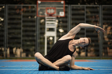 Happy fitness sporty man in black sportswear stretching his arms and warming up while sitting on the floor on outdoor basketball court. Beautiful man doing work out and different exercises outdoor