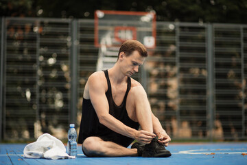Fototapeta na wymiar Sporty muscular man tying laces on sport sneakers before morning run at outdoor basketball court. Young fitness man training on fresh air.