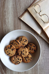 Plate of chocolate chip cookies, open books and reading glasses on wooden table. Flat lay.