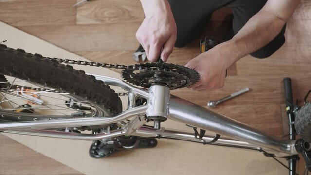 Man removes front chainring of inverted bicycle with connecting rod and pedals from aluminum frame of vehicle, top view. Mechanic removes chain from sprockets. Dismantling bike for repair.