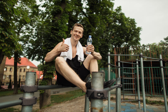 Little break. Young athletic guy with muscular body, sportsman and fitness model, strong man sitting on parallel bars. Athletic body shape, guy relax at outdoor gym showing thumb up.