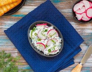 Appetizer, salted cottage cheese with dill, green onion and radish in a black bowl on a wooden background. Recipes with cottage cheese.