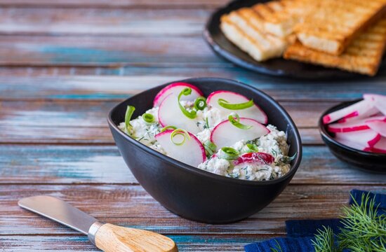 Appetizer, salted cottage cheese with dill, green onion and radish in a black bowl on a wooden background. Recipes with cottage cheese.
