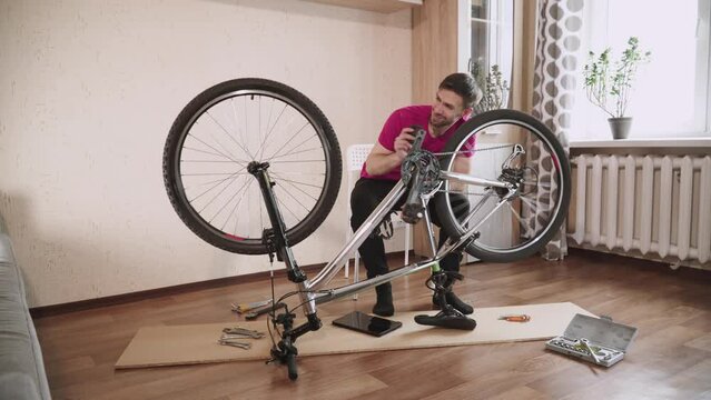 Man turns the pedals inverted bicycle with hand in residential apartment. Guy listens to sound, wheel squeaks when spinning. Upset cyclist shakes head in displeasure and leans back in his chair.