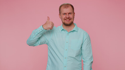 Bearded adult man in casual blue shirt raises thumbs up agrees with something or gives positive reply recommends advertisement likes good. Young casual guy isolated alone on pink studio background