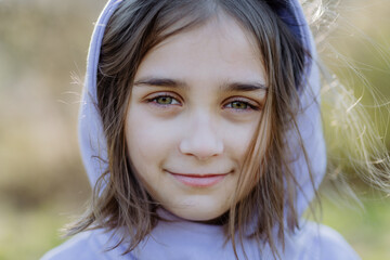 A portrait of pretty child girlin hoodie standing in summer park looking at camera smiling happily.