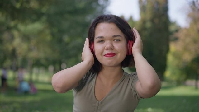 Portrait of relaxed Caucasian young woman with dwarfism listening to music on headphones smiling dancing in slow motion. Happy confident carefree little person looking at camera posing in park