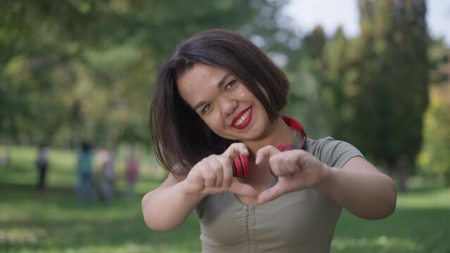Portrait of positive little woman gesturing heart shape looking at camera smiling sitting in summer park outdoors. Happy confident Caucasian person with dwarfism posing in slow motion resting