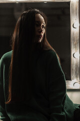 Beautiful Asian girl in a vintage green sweater sits in the dark near the mirror with light bulbs