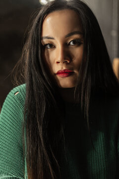Female portrait of a pretty young stylish Vietnamese woman with makeup in a fashionable vintage knit sweater looks at the camera in a room in the dark