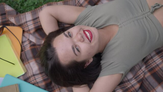 Top view portrait of happy Caucasian little woman looking at camera smiling lying on blanket outdoors. Relaxed carefree person with dwarfism enjoying leisure in spring summer park posing