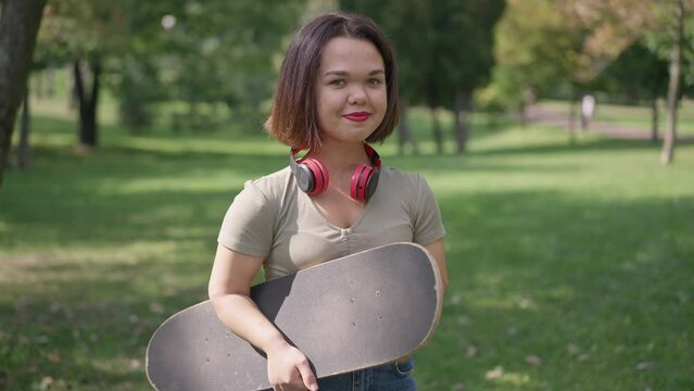 Medium shot portrait of charming young woman with dwarfism posing in park with skateboard. Smiling happy Caucasian little person looking at camera standing outdoors on spring summer day