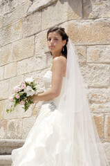 Beautiful girl dressed in a wedding dress and with a bouquet of flowers next to a stone wall looking at the camera on her sunny wedding day.