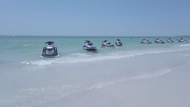 A row of wave runners are seen bobbing in beautiful clear ocean water near a soft white beach 