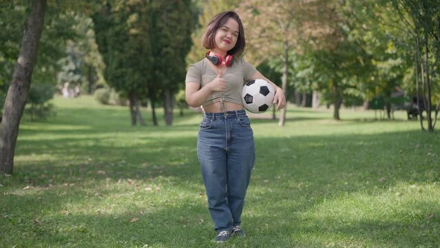 Wide shot fit young woman with dwarfism gesturing thumb up smiling looking at camera standing with football ball in summer park. Front view positive little person posing in slow motion