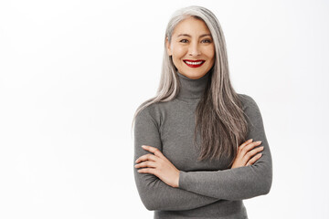 Portrait of beautiful, healthy smiling middle aged asian woman, cross arms on chest, looking confident and happy, standing over white background