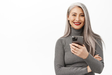 Portrait of beautiful middle aged asian woman holding smartphone and smiling at camera, using mobile app, standing over white background