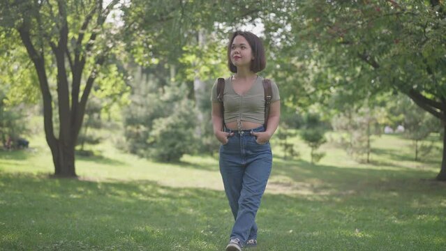 Wide shot happy smiling young woman with dwarfism walking in sunny forest in slow motion. Portrait of relaxed positive little person travelling in woodland outdoors strolling