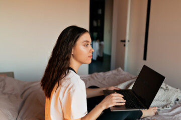 Close up photo of brunette lovely woman with dark wavy hair working on laptop and looking aside with smile in bedroom