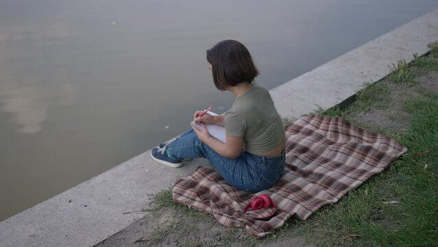 High angle view of thoughtful Caucasian little woman writing diary sitting on lake shore in summer park. Wide shot happy relaxed young person with dwarfism dreaming resting outdoors