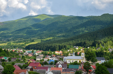 Fototapeta na wymiar Beautiful rural landscape with colorful houses and church in the green meadow at the foot of the mountain ridge covered forest. Kolochava village in Carpathian Mountains, Transcarpathian, Ukraine
