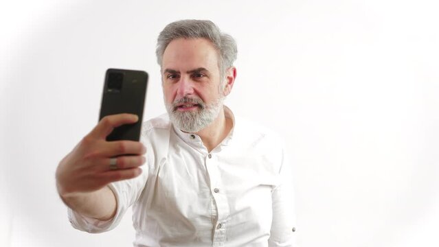 Funny elder gray-haired entrepreneur man taking selfie with his black smartphone and doing silly faces over white background. High quality 4k footage