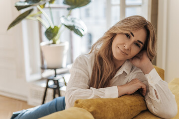 Obraz premium Beautiful middle-aged caucasian woman looking at camera while sitting on comfortable yellow sofa at home. Blonde wears white blouse while relaxing alone. Rest time concept.
