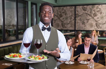 Hospitable African American waiter standing with serving tray, recommending dishes in restaurant