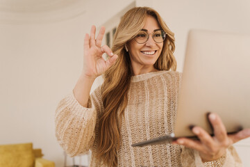 Cheerful adult caucasian lady smiles, shows ok sign in notebook screen on white background. Successful blonde woman in glasses leads video call. Technology concept