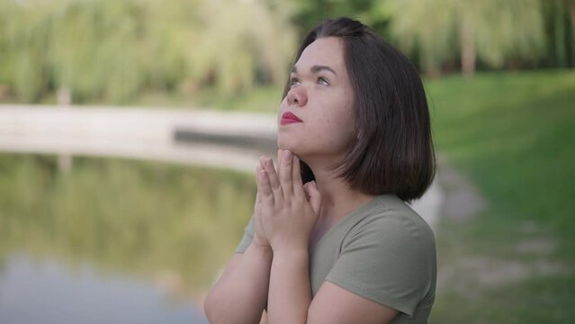 Side view hoping young little woman praying standing in summer park on lake shore. Portrait of smiling positive Caucasian young person with dwarfism thanking God standing outdoors