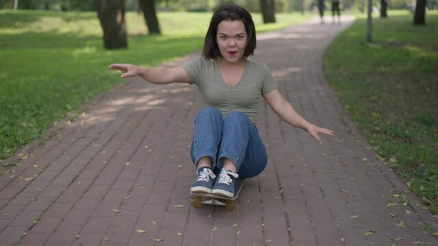 Dolly shot of joyful little person sitting on skateboard riding in summer park on alley. Wide shot portrait of happy Caucasian woman with dwarfism having fun on weekend outdoors