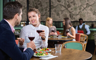 Attractive cheerful smiling girl with boyfriend enjoying dinner with wine in cozy restaurant