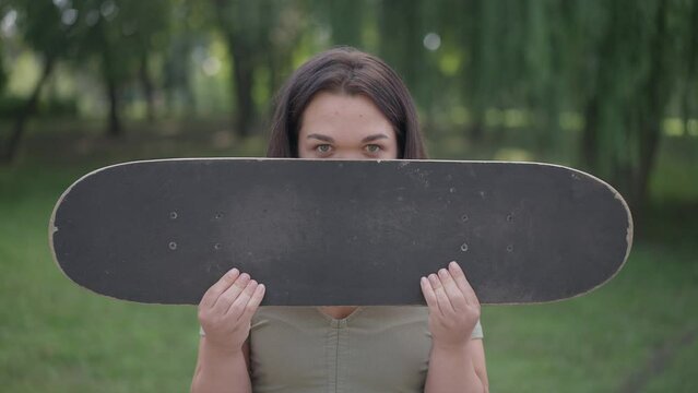 Cheerful Caucasian little person hiding face with skateboard looking at camera smiling. Front view portrait of joyful young woman with dwarfism posing in slow motion in summer park