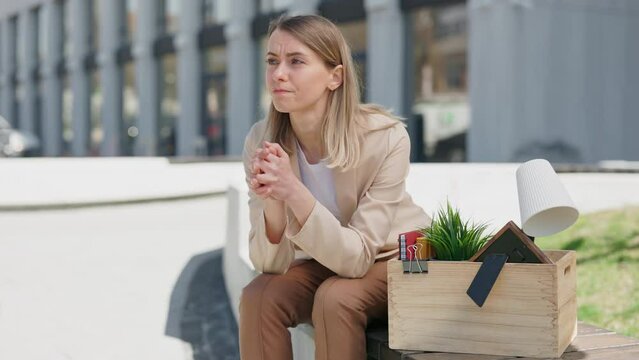 Frustrated young caucasian woman sitting on bench with box of personal belongings near business center. Sad female worker fired from large corporation.