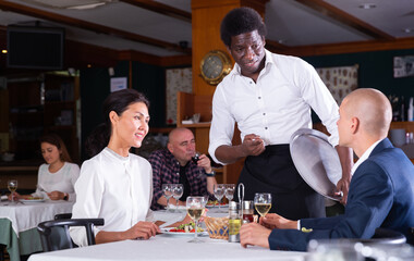 Fototapeta na wymiar smiling male waiter serving order to adult man and woman in restaurante