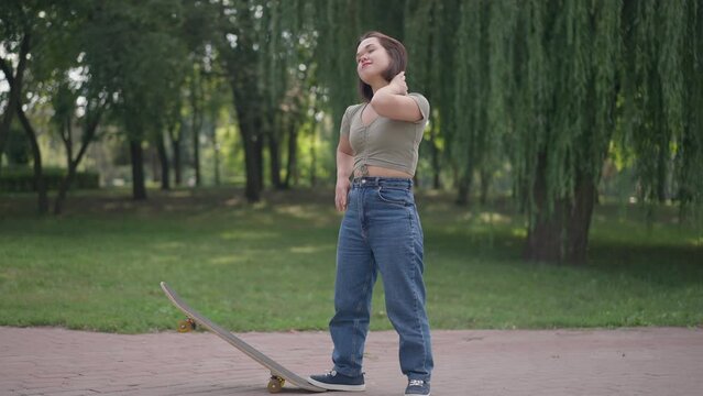 Wide shot confident Caucasian young woman with dwarfism standing with skateboard in spring summer park smiling looking away. Happy positive fit little person enjoying weekend leisure outdoors
