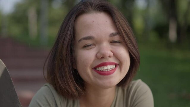 Front view close-up charming little woman smiling looking at camera sitting with skateboard in summer park. Portrait of happy confident fit Caucasian person with dwarfism posing outdoors