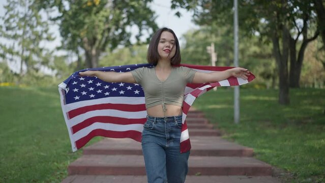 Confident proud little woman with dwarfism walking in slow motion holding American flag looking at camera. front view portrait of smiling Caucasian little person posing strolling in park outdoors