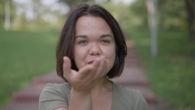 Close-up coquette Caucasian little woman gesturing air kiss smiling looking at camera. Portrait of confident happy positive young person with dwarfism posing in slow motion flirting standing in park