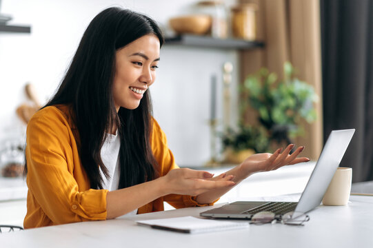 Friendly pretty asian brunette girl, manager, freelancer, sit in the kitchen, uses laptop, talking by video call with the client or employees, discussing project, holds consultation, smiling
