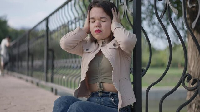 Nervous stressed little person with dwarfism having migraine holding head in hands closing eyes. Portrait of overburdened frustrated Caucasian young woman with headache in park outdoors